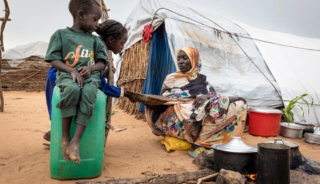 In a displacement camp in Sudan, a woman cooks over an open fire outside a makeshift shelter as two children watch. Millions have been displaced over the past two years of conflict. In a displacement camp in Sudan, a woman cooks over an open fire outside a makeshift shelter as two children watch.